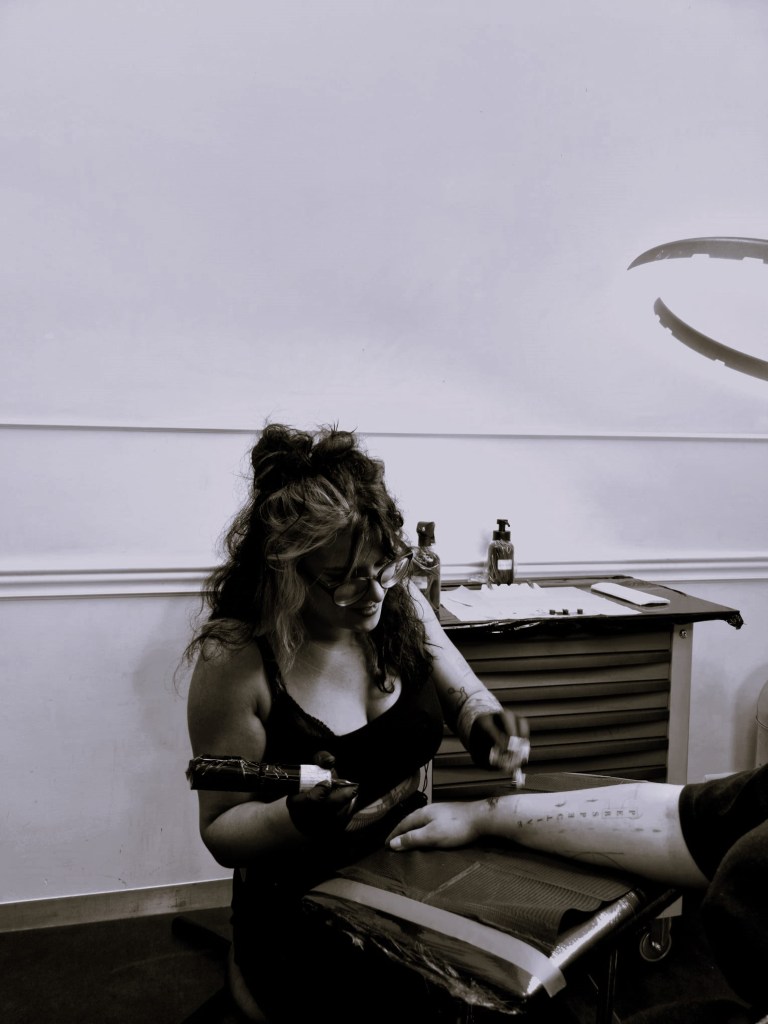 Tattoo Mercé working on a client's arm in a professional studio, with tattoo tools and supplies visible on a nearby cart.