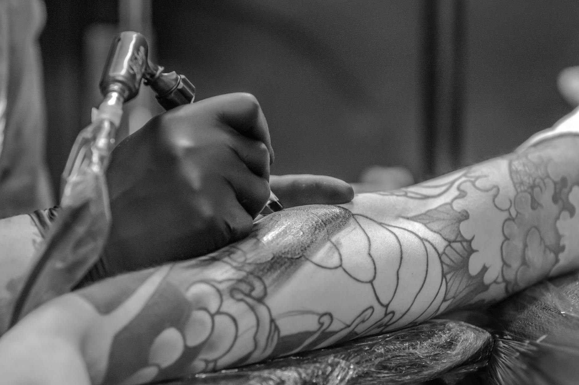 Close-up of a tattoo artist shading a floral tattoo design on a client’s arm using a tattoo machine. The intricate linework and shading detail stand out in the monochrome setting.